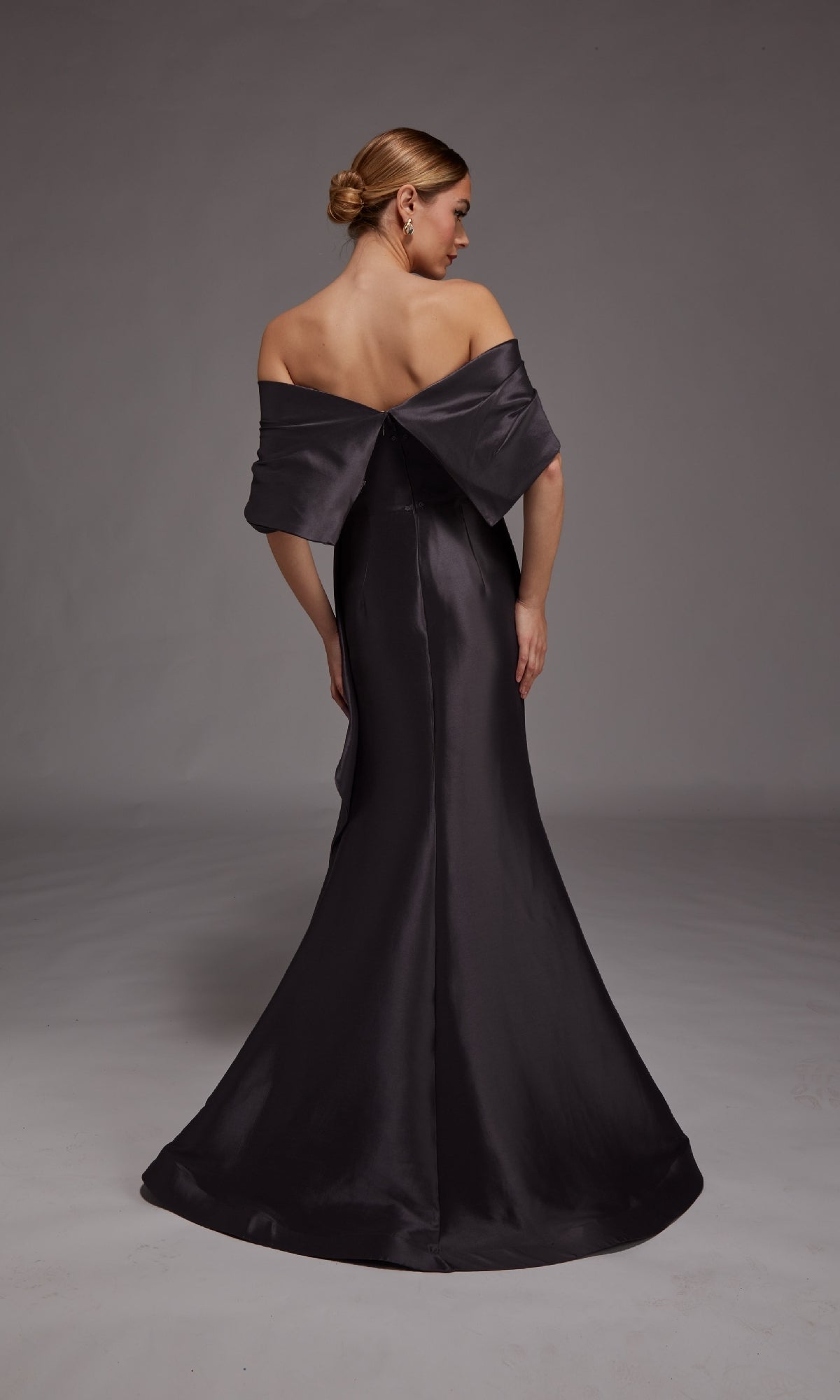 A woman with her hair in a low bun stands facing away from the camera against a gray background, wearing the Alyce 27822 Long Formal Dress—an off-the-shoulder, floor-length gown with a mermaid silhouette.